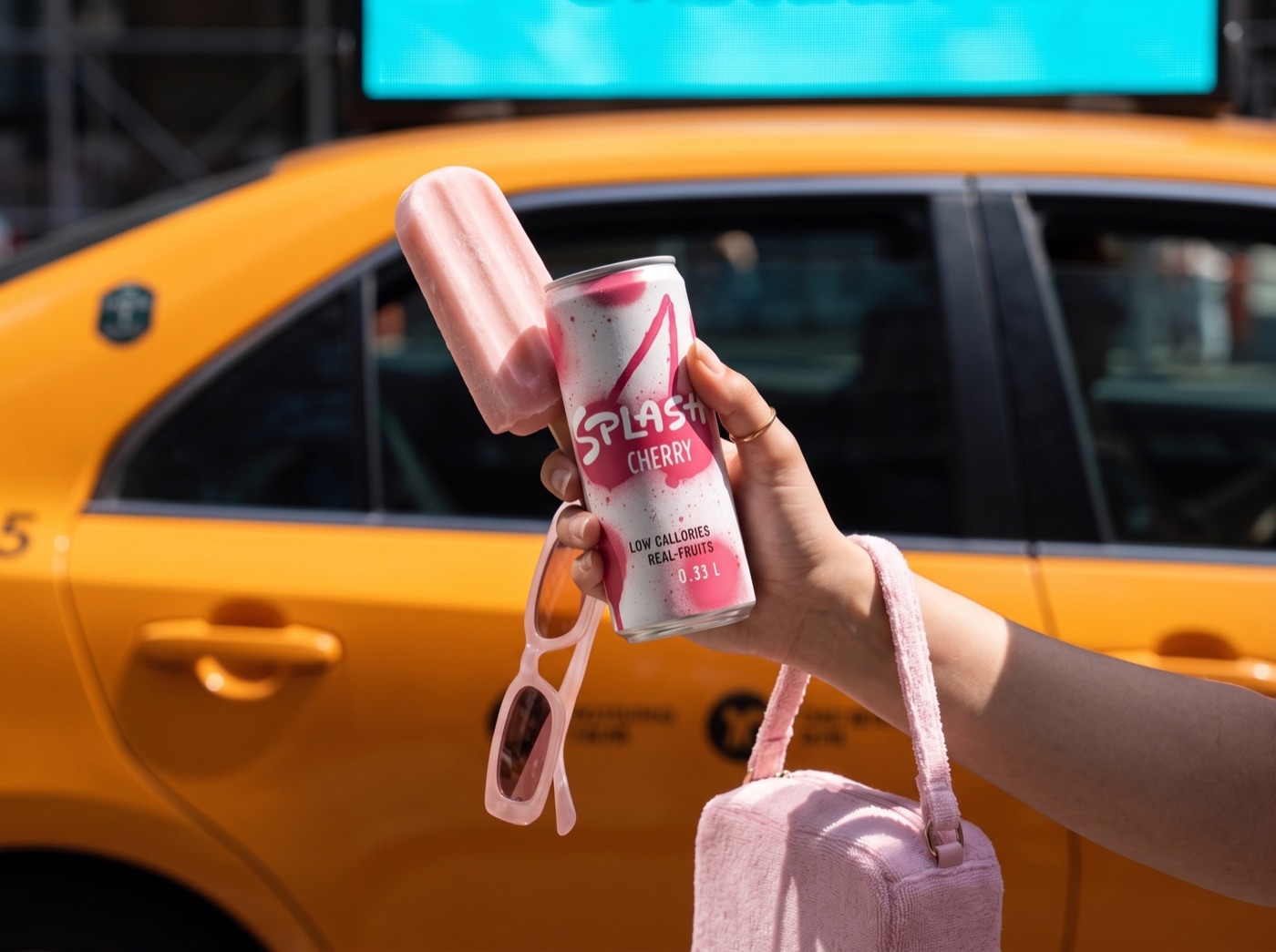 Hand holding a cherry drink can and an ice lolly in front of a yellow taxi